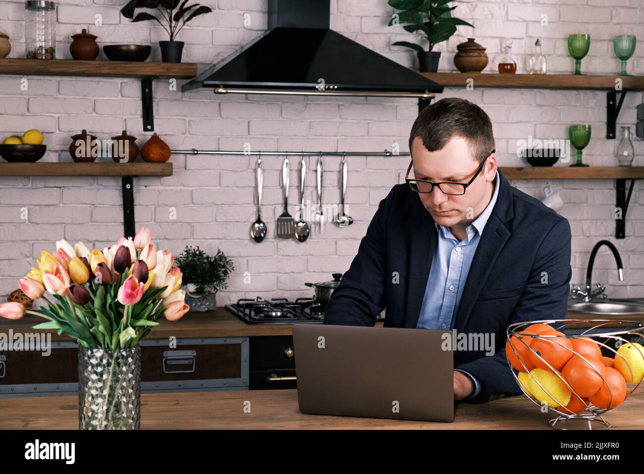 Lavoro in remoto con il computer portatile. Ritratto di giovane uomo d'affari serio in occhiali e tuta da lavoro, seduto a casa in cucina con computer portatile e guardando Foto Stock