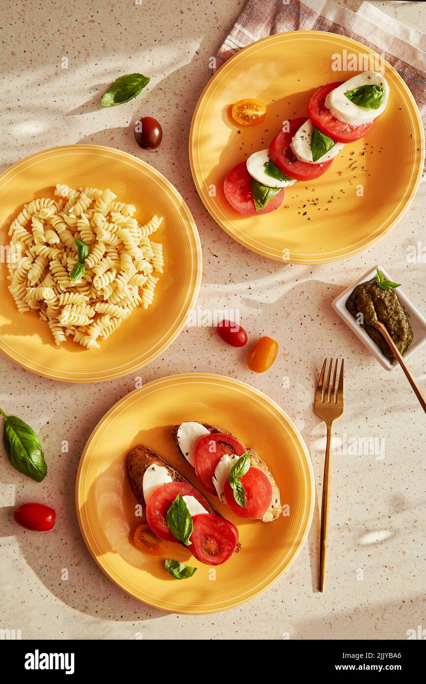 Pasta fusilli mediterranea, insalata caprese italiana - pomodori, mozzarella, basilico e pesto. Antipasto di pane con insalata caprese su piatti d'arancia sotto ombre alla moda. Vista dall'alto cibo piatto Foto Stock