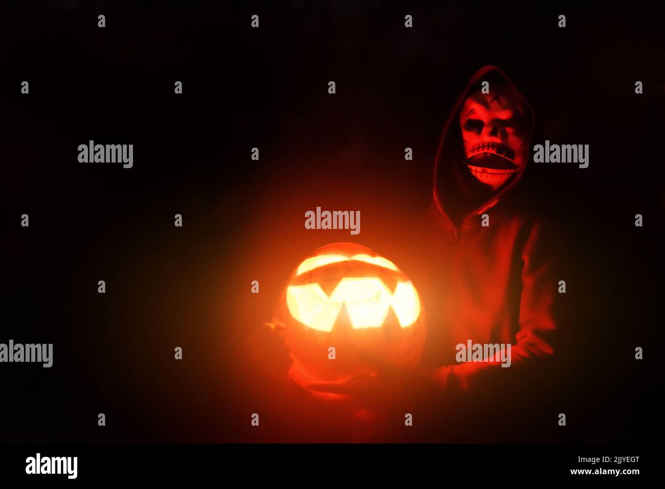 Terribile uomo mascherato illuminato con luce rossa da lampada di zucca di Halloween si trova vicino alla casa di notte. Concetto di Halloween Foto Stock