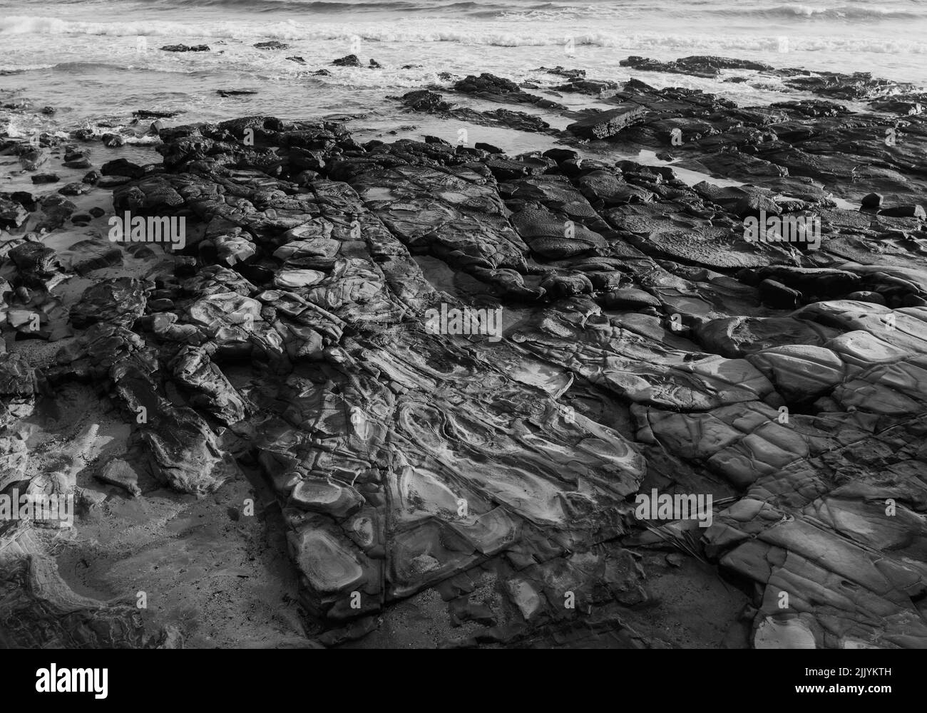 Le onde si lavano dolcemente su formazioni rocciose sulla sabbia - B&W. Foto Stock