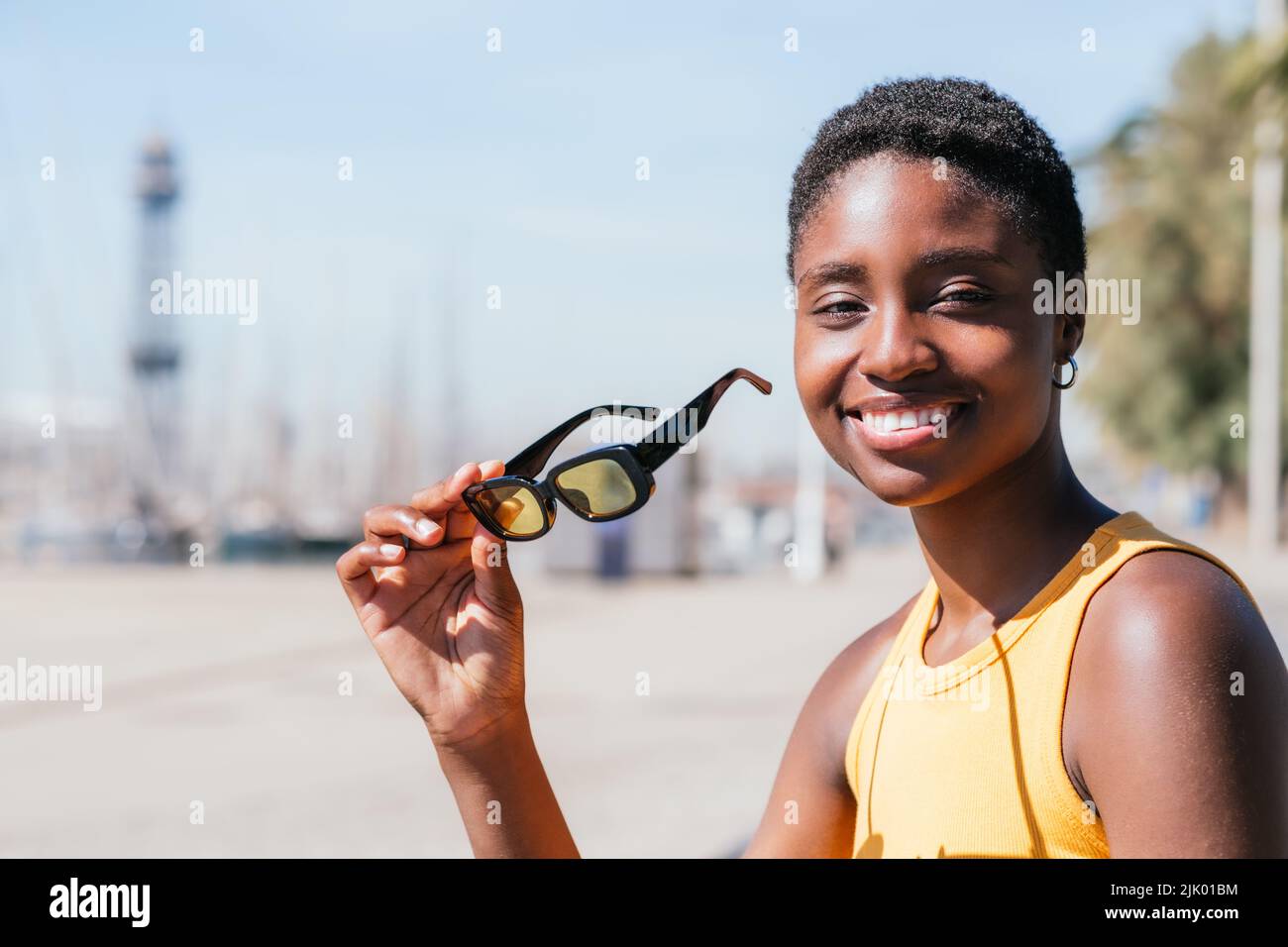 ritratto di una donna afro-americana positiva guardando la macchina fotografica vicino al porto. Indossa una t-shirt di colore giallo estate e tiene occhiali da sole gialli Foto Stock