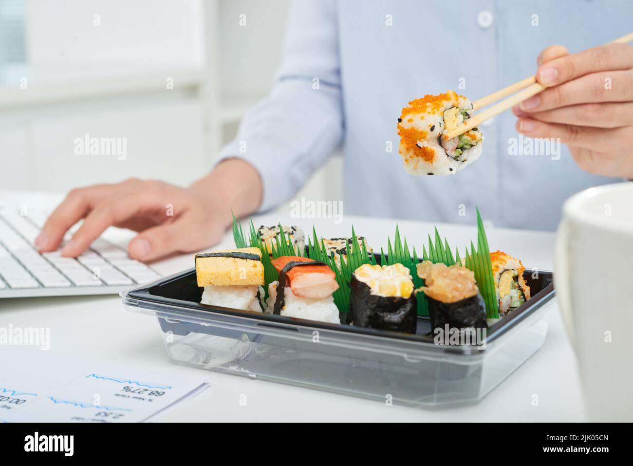 Primo piano di un operaio di colletto bianco occupato seduto di fronte al computer e mangiare sushi con l'aiuto di bacchette Foto Stock