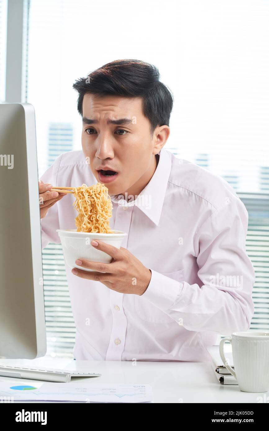 Scioccato colletto bianco asiatico che guarda lo schermo del computer mentre si mangia spaghetti istantanei con bacchette, vita-up ritratto girato Foto Stock