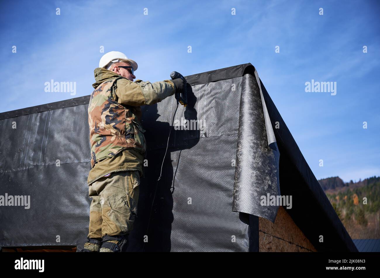 Maschio costruttore installare membrana impermeabile sul muro del futuro cottage. Uomo lavoratore edificio legno telaio casa con cielo blu sullo sfondo. Carpenteria e concetto di costruzione. Foto Stock