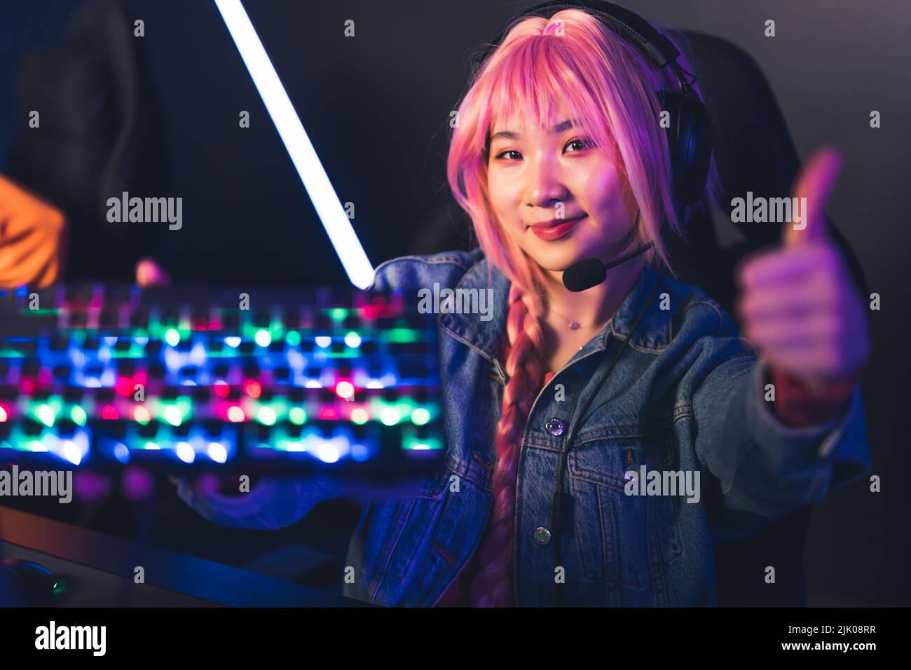 Ragazza asiatica adolescente con rosa sentire indossare cuffie tenere la tastiera di gioco incandescente colorato fino alla fotocamera e dare i pollici in su. Foto di alta qualità Foto Stock