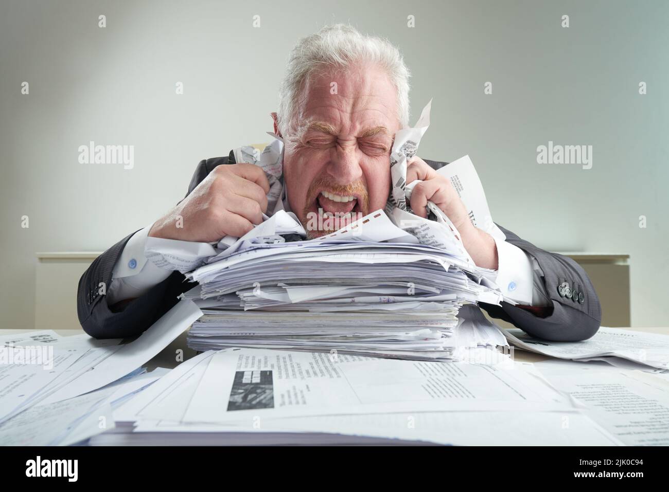 Ritratto di impazzito imprenditore senior poggiando il mento su un mucchio di documenti, urlando e crumpling liste di carta mentre alleviando lo stress, ufficio interno su sfondo Foto Stock