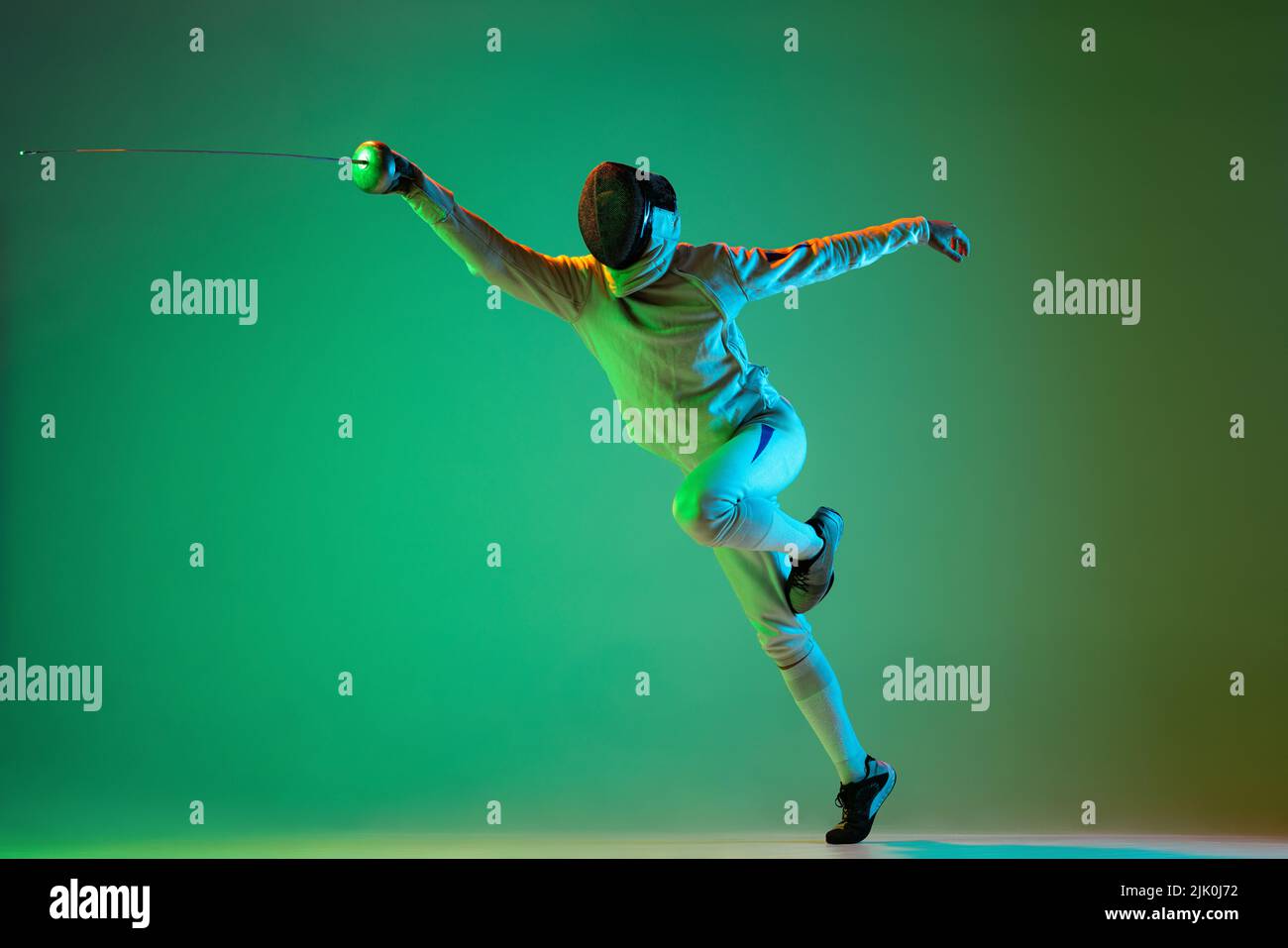 Giovane uomo energico, fencer con spada di piccolo pratica scherma isolato su sfondo verde in luce al neon. Sport, energia, abilità, realizzazioni Foto Stock