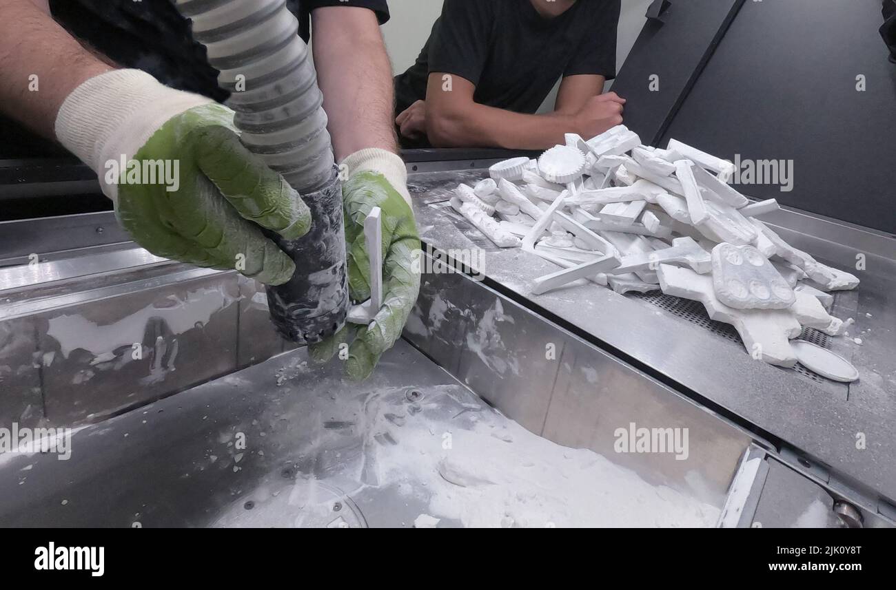 Un uomo che lavora con un aspirapolvere funzionante per pulire la polvere bianca di poliammide da un modello stampato su una stampante 3D all'interno di una stampante 3D. Pulizia di oggetti stampati su una stampante industriale a polvere 3D Foto Stock