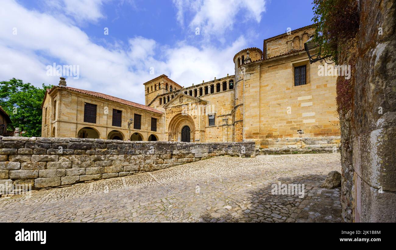 Panoramica della vecchia chiesa romanica in pietra con pavimentazione in ciottoli e cielo blu. Santillana del Mar. Foto Stock