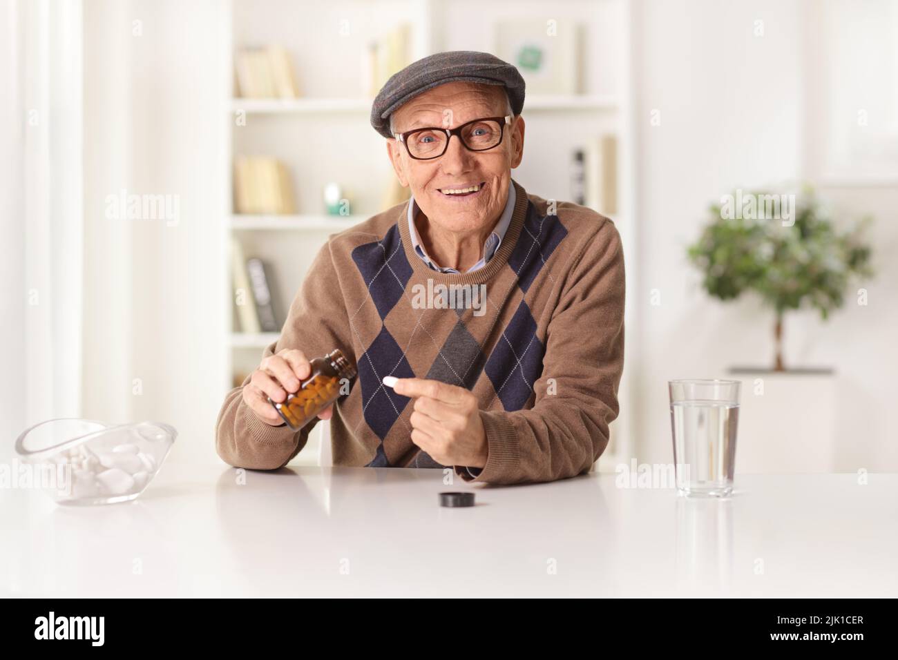 Uomo anziano sorridente seduto a casa e prendendo una pillola da una bottiglia Foto Stock