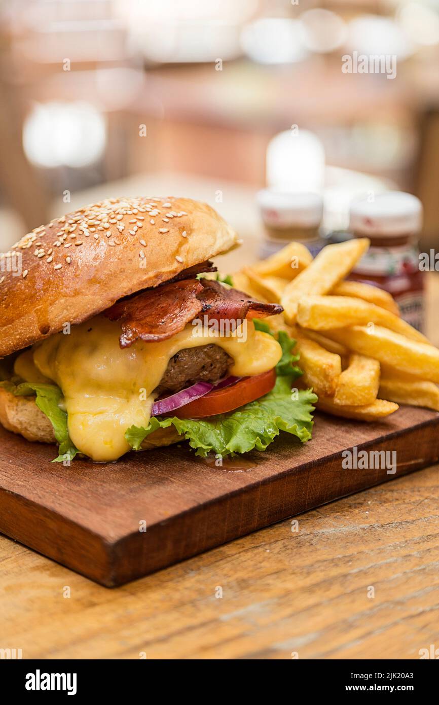 Cheeseburger con formaggio, lattuga, pomodoro, cipolla, pancetta e patatine fritte, servito su tavola di legno in un ristorante durante il giorno Foto Stock