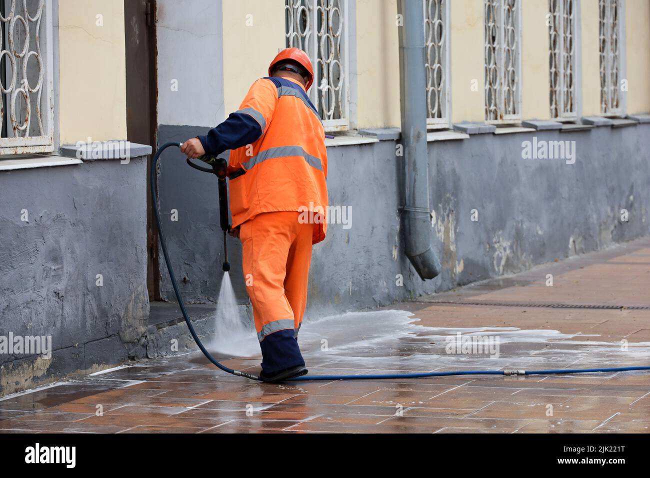 L'operatore innaffia il marciapiede con un tubo flessibile. Pulizia e disinfezione della strada nella città estiva Foto Stock