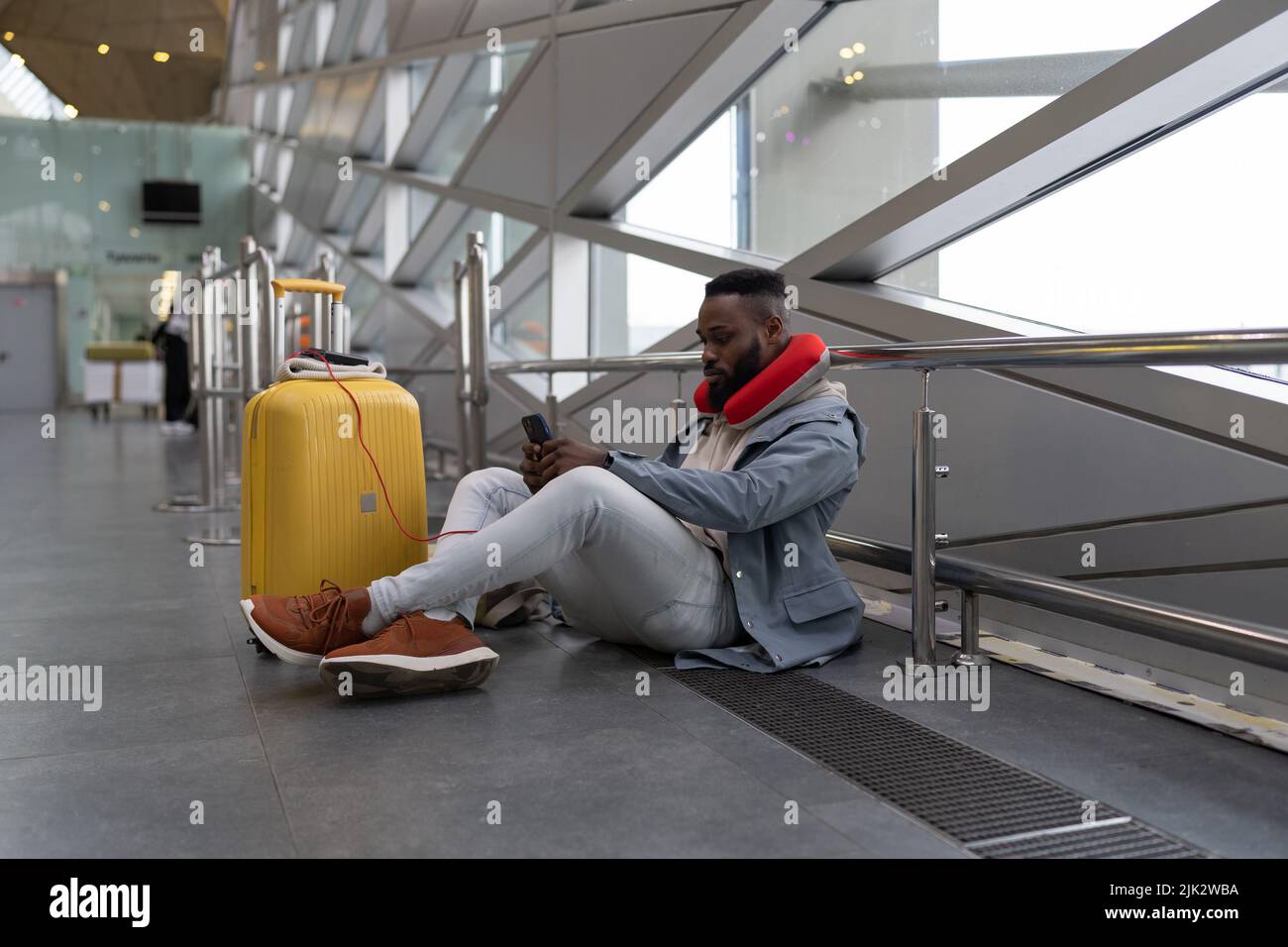 Il giovane uomo nero serio si siede a gambe incrociate sul pavimento in attesa di volo utilizzando power bank per cellulare Foto Stock