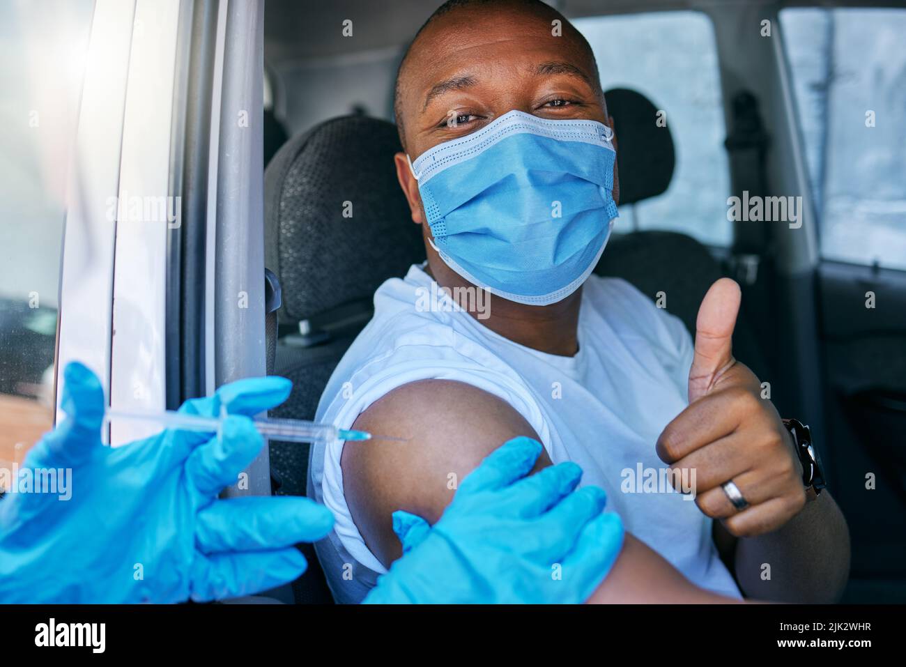 Guidare attraverso il sito di vaccino del virus della corona e del covid come servizio pubblico per la gente. L'uomo che dà i pollici in su ed endorsing il jab mentre porta una maschera a. Foto Stock