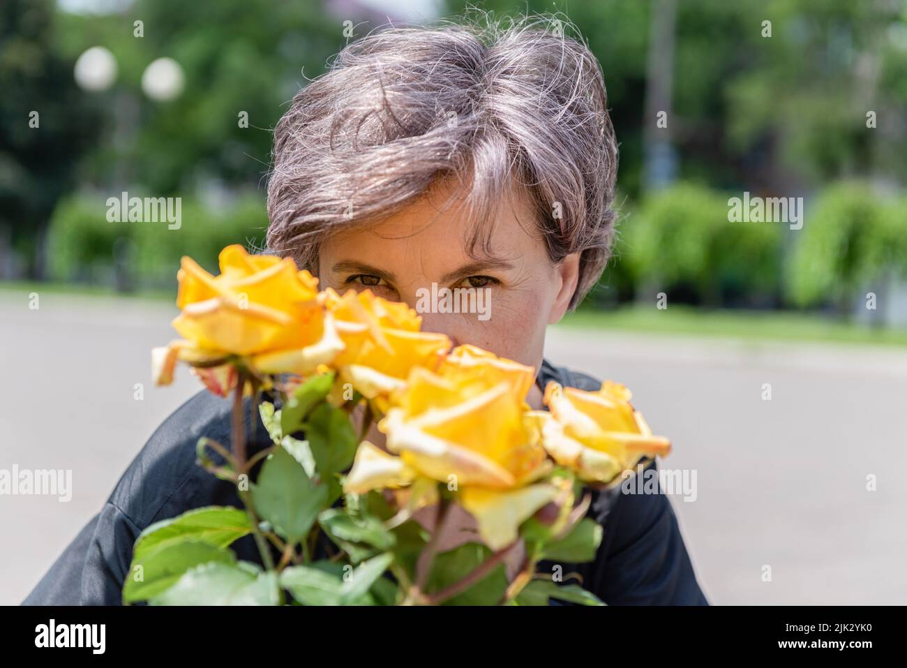 Donna che sbircia da dietro un bouquet di rose gialle in fiore contro uno sfondo sfocato del parco. Foto Stock