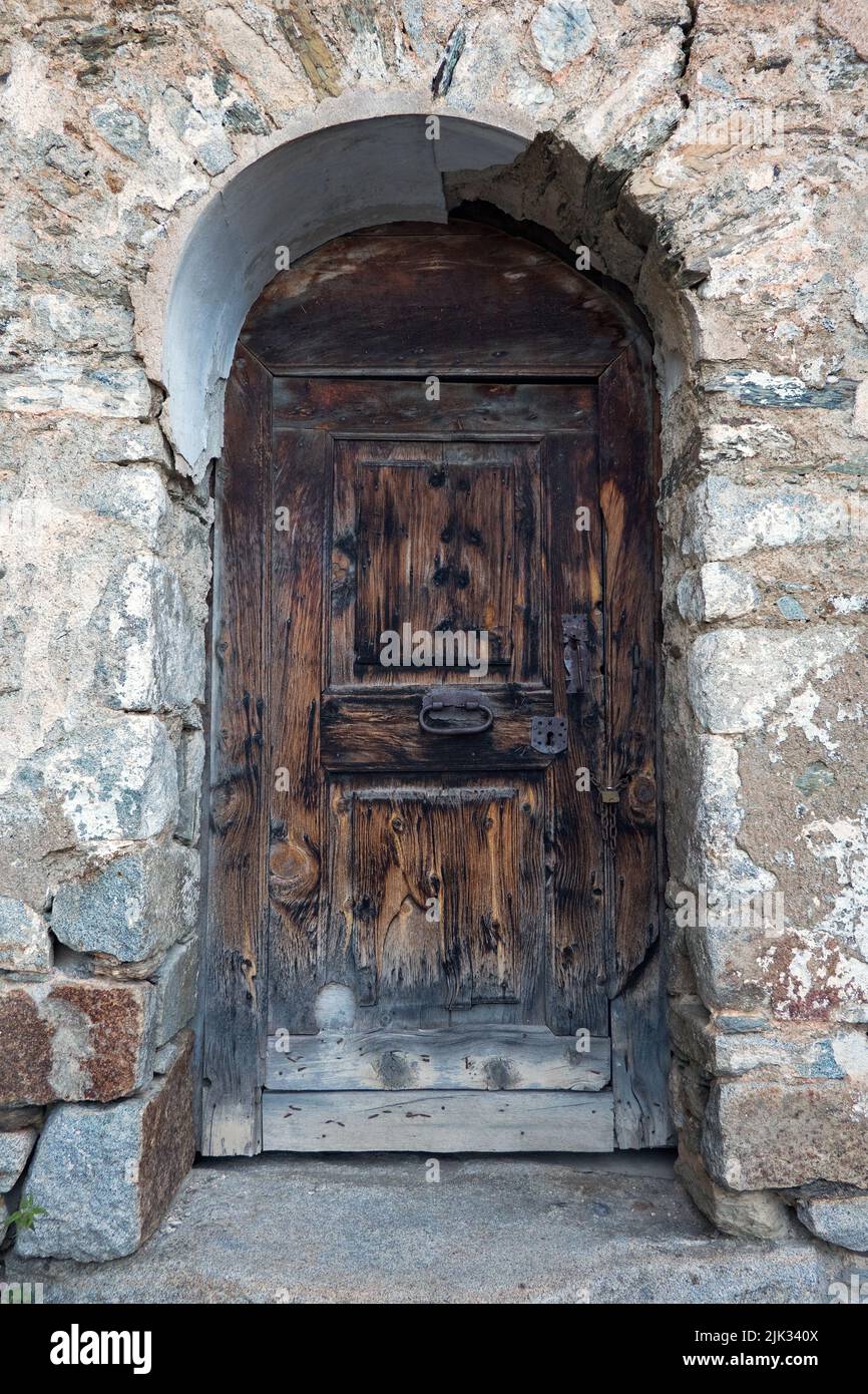 Vecchia porta in legno intemperie con piastre di bloccaggio arrugginite e maniglia in un muro, mascherata con grandi pietre naturali Foto Stock