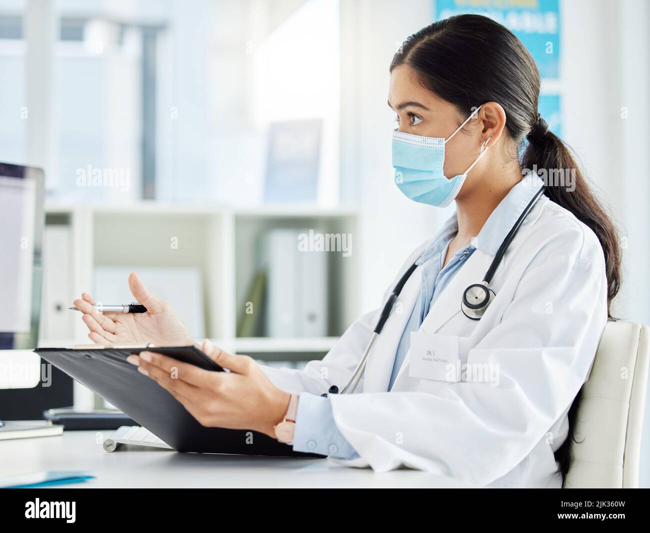 Avete domande: Un medico che indossa una maschera mentre si siede e parla nella sua clinica durante una consultazione. Foto Stock