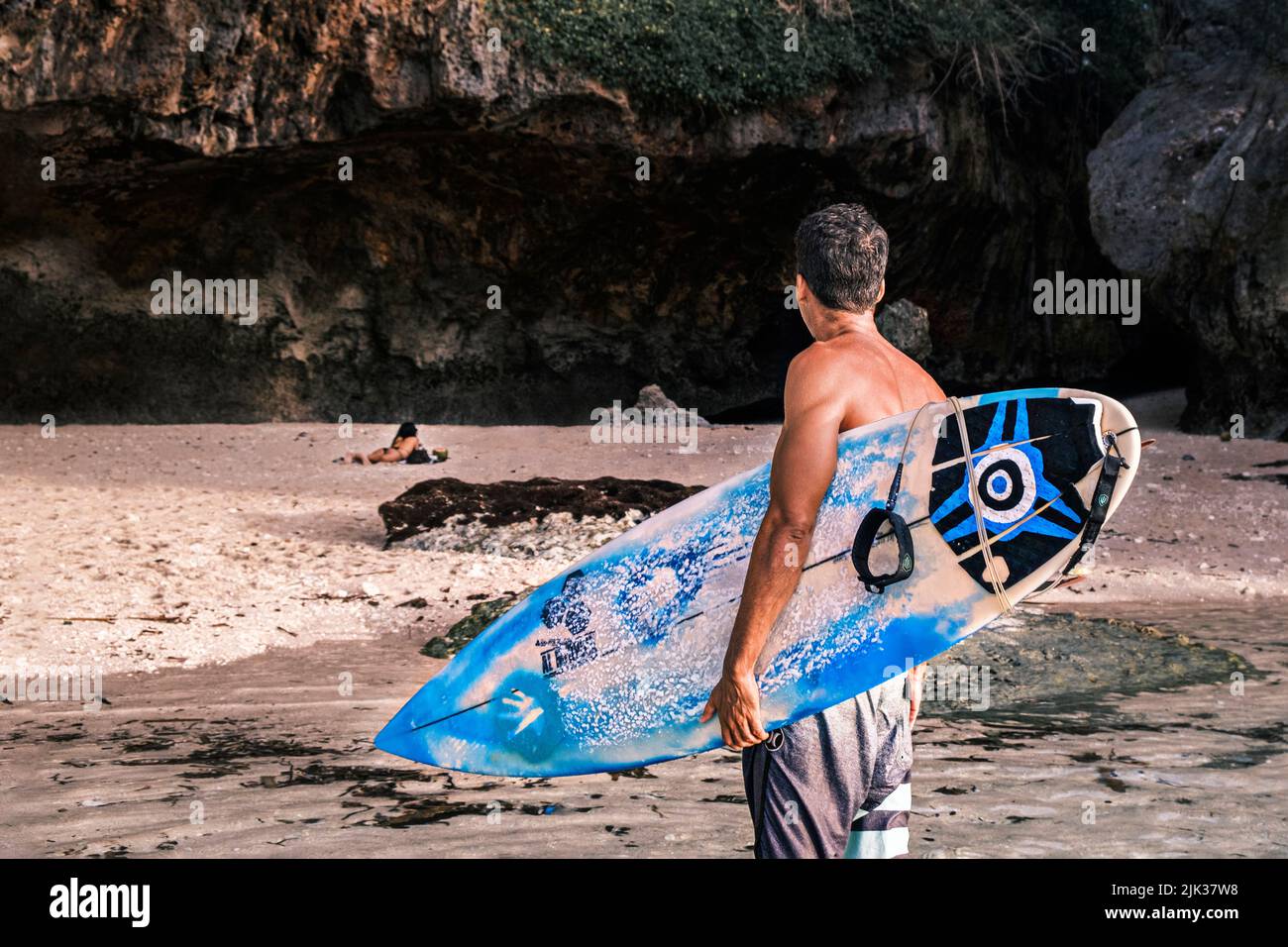 Vista posteriore di professionista surfista nero tenendo la sua tavola da surf in mano si alza su una spiaggia oceano contro la scogliera guardando la giovane donna prendere il sole Foto Stock