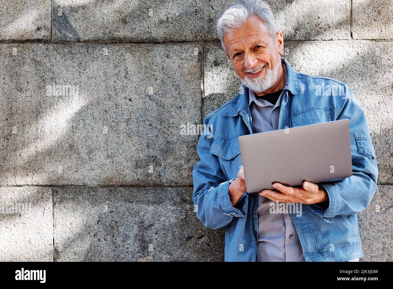 Uno studente anziano felice sta in piedi contro il muro di mattoni e sta scrivendo su un notebook. Foto Stock