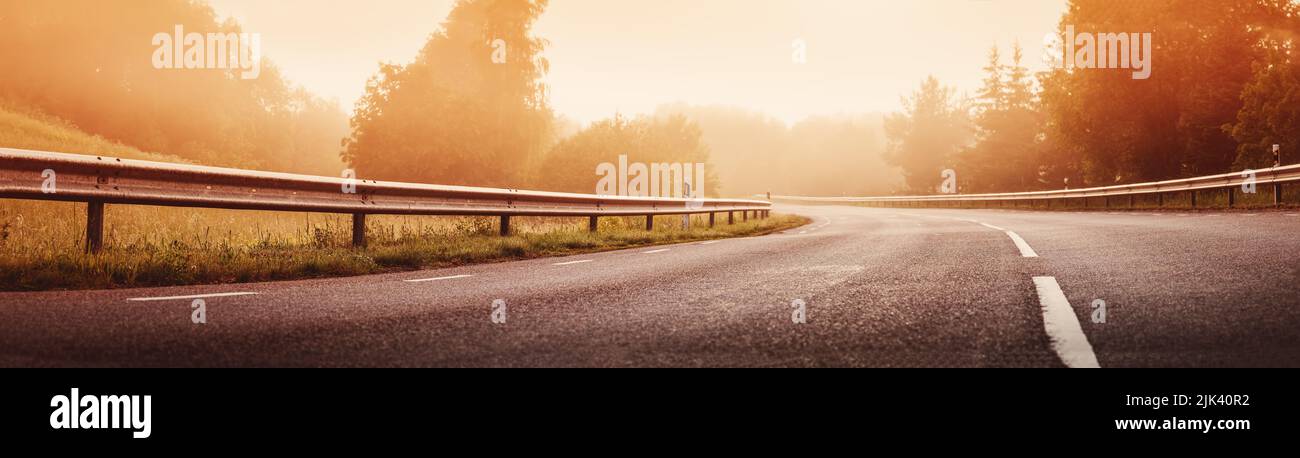 Strada asfaltata nera con linea di demarcazione e recinzione di sicurezza nella nebbia mattutina. Foto Stock