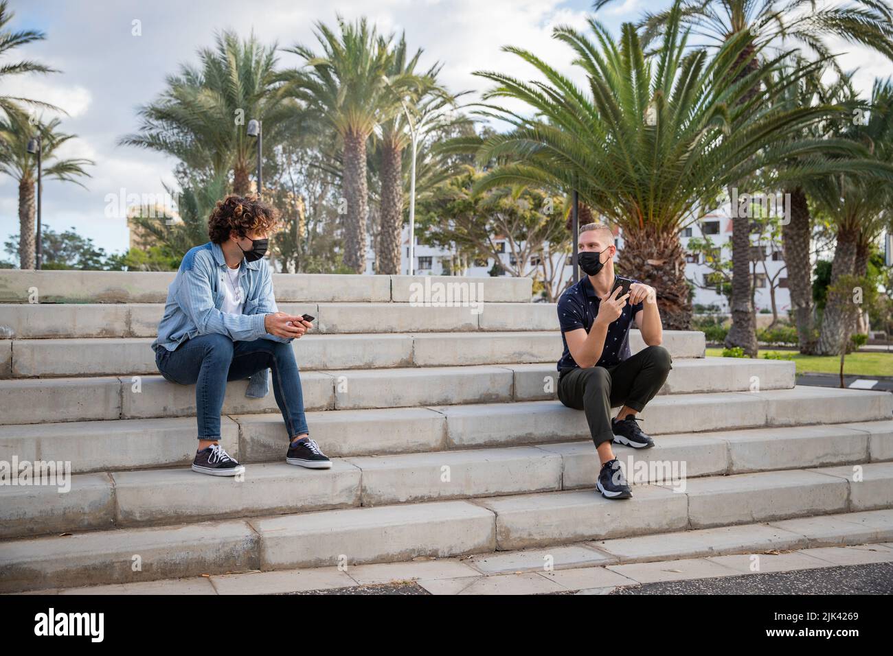 Due ragazzi seduti sulle scale all'esterno utilizzano gli smartphone indossando maschere chirurgiche e mantenendo una distanza di sicurezza durante la pandemia del coronavirus Foto Stock