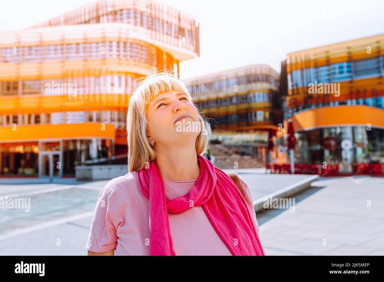 donna bionda di mezza età 40s con capelli lunghi, in sciarpa rosa e abiti estivi, guarda all'architettura moderna con speranza e sognamento. concetto di husi Foto Stock