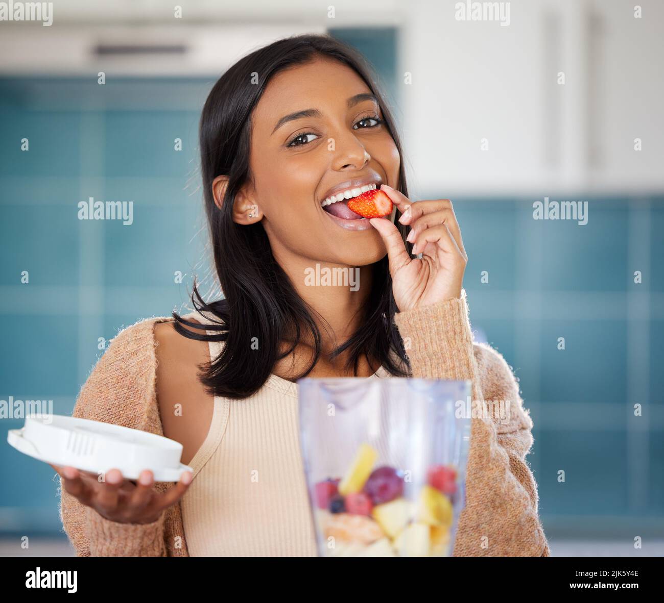 Scegli gli alimenti che nutrono il tuo corpo. Una giovane donna che mangia una fragola mentre prepara un frullato sano a casa. Foto Stock Scegli gli alimenti che nutrono il tuo corpo. Una giovane donna che mangia una fragola mentre prepara un frullato sano a casa. Foto Stock