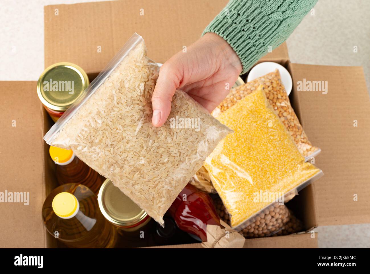 Contenitore in plastica con riso crudo in mano femmina sullo sfondo della scatola per alimenti di emergenza Foto Stock