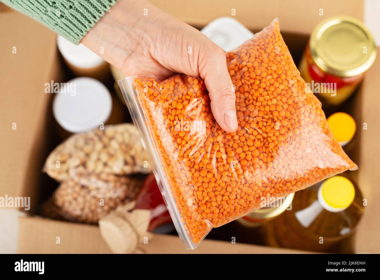 Contenitore in plastica con lenticchie in mano femmina sullo sfondo della scatola per alimenti di emergenza Foto Stock