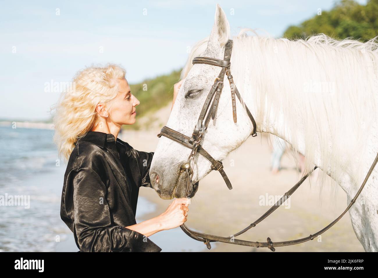 Giovane donna bionda in abiti neri a cavallo bianco su sfondo marino Foto Stock