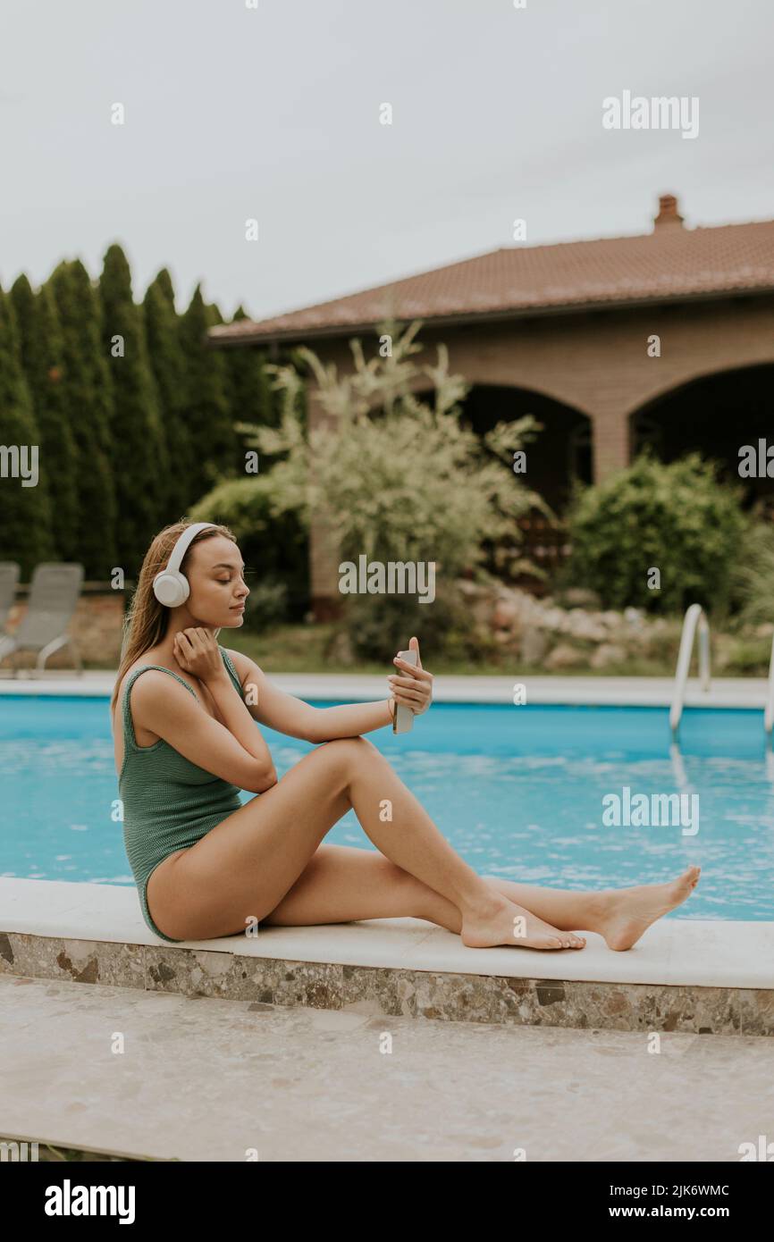 Bella giovane donna seduta in piscina e ascoltare musica dal telefono cellulare con cuffie nel cortile della casa Foto Stock
