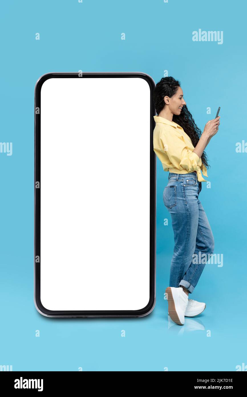 Donna che utilizza smartphone appoggiandosi su un enorme schermo del telefono, sfondo blu Foto Stock