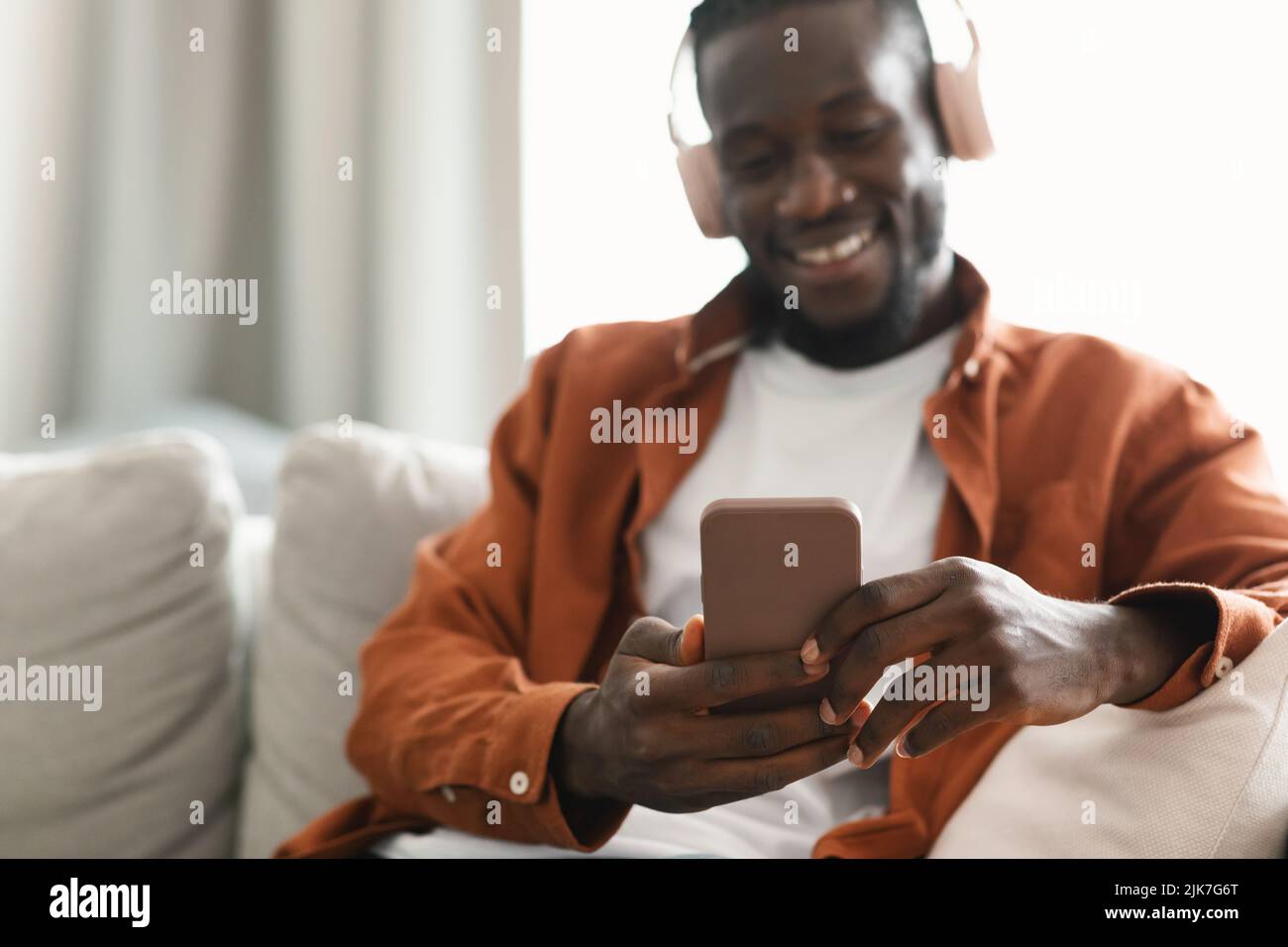 Eccitati afroamericani che navigano su internet mentre si riposano sul divano di casa, utilizzando cuffie wireless e telefono cellulare Foto Stock