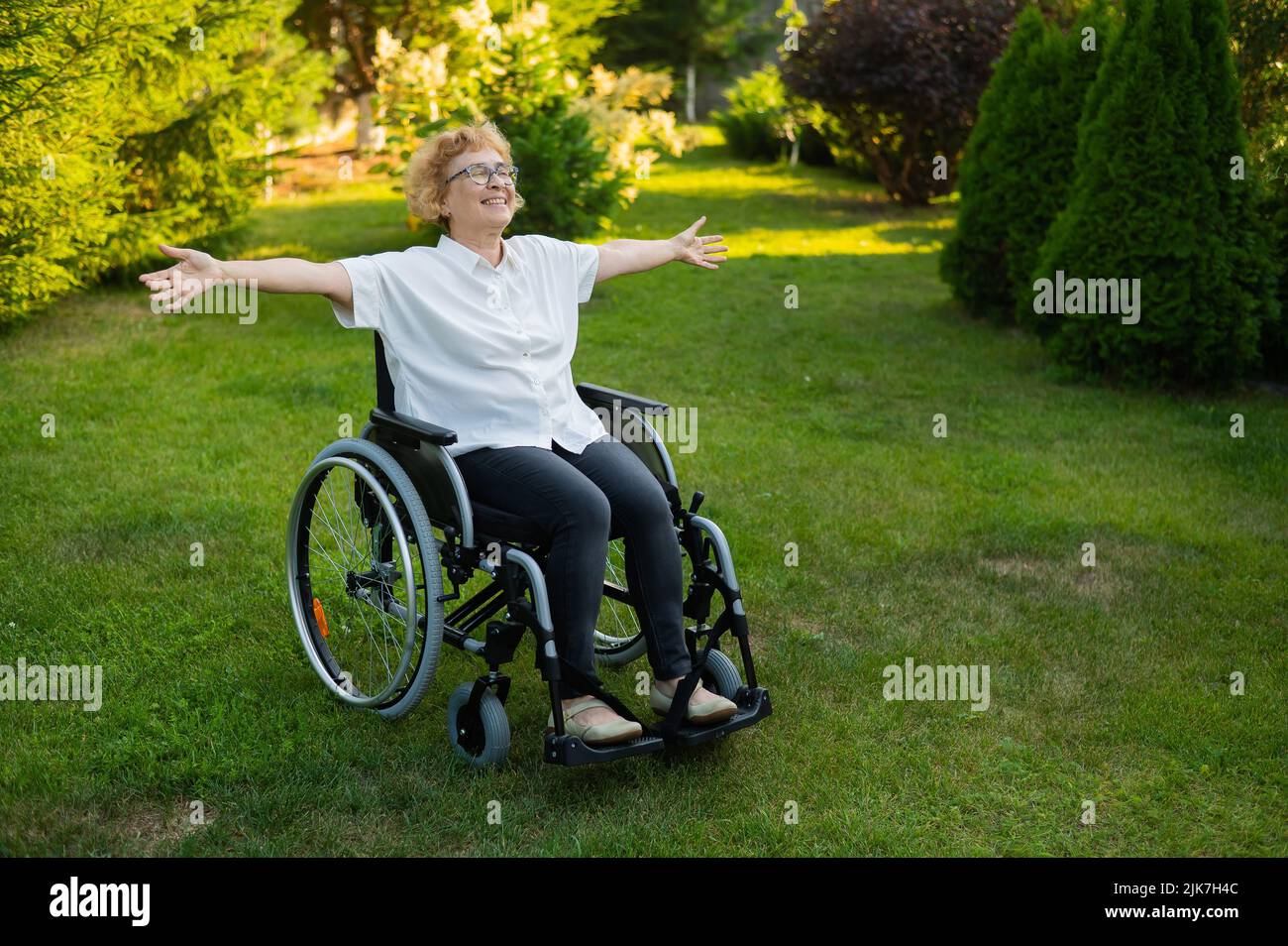 La donna anziana felice si stese le braccia ai lati mentre si siede in una sedia a rotelle su una passeggiata all'aperto. Foto Stock