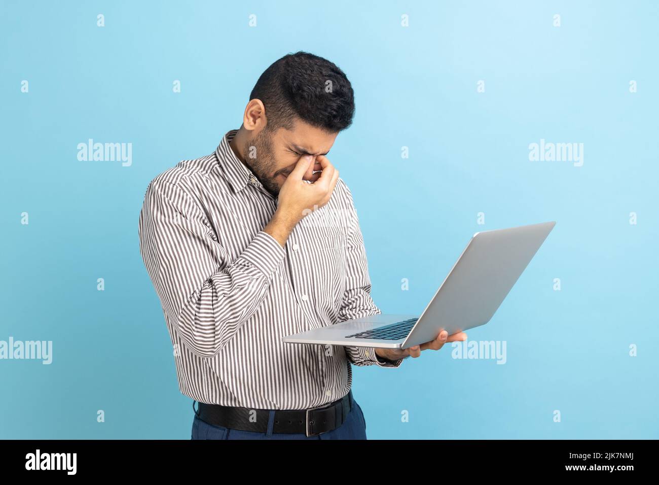Ritratto di giovane uomo d'affari adulto in piedi con il laptop in mani, strofinando gli occhi, sembra stanco e esausto, indossando una camicia a righe. Studio interno girato isolato su sfondo blu. Foto Stock