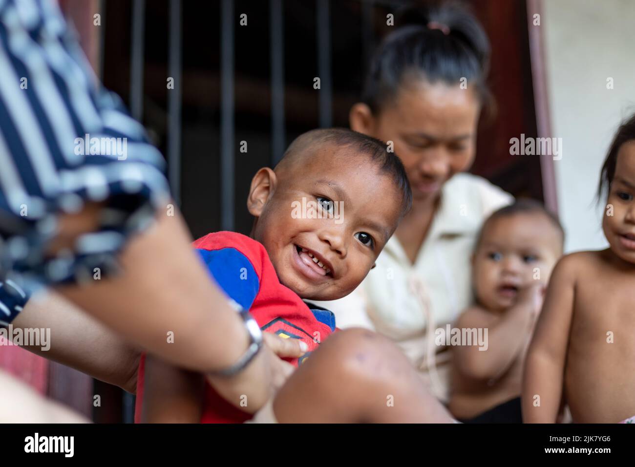 Un giovane ragazzo thailandese sorride mentre è solletico dal suo sponsor, mentre sua madre e le sue sorelle si siedono dietro in uno sfondo sfocato Foto Stock