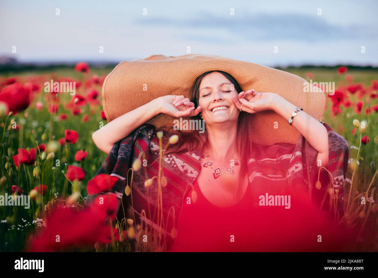 Ritratto di una donna sorridente in un campo di papavero che indossa un cappello a larga sposa al tramonto. Vista frontale. Foto Stock
