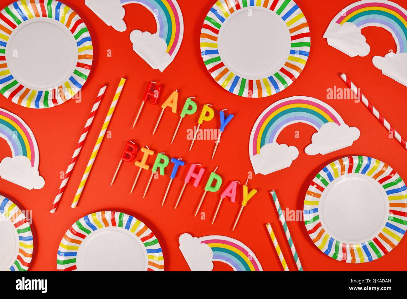 Festa di compleanno piatto con piatti colorati, tovaglioli arcobaleno, candele e cannucce da bere su sfondo rosso Foto Stock