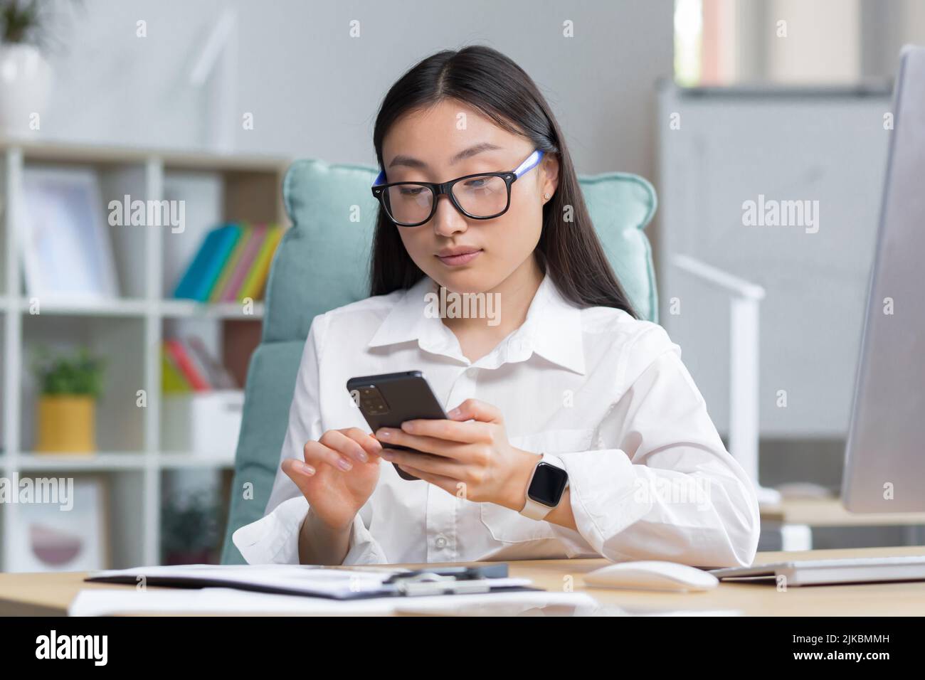 Una giovane bella donna asiatica d'affari in occhiali sta tenendo un telefono nelle sue mani, componendo un messaggio. Seduto su una scrivania in un ufficio moderno, guardando la fotocamera Foto Stock