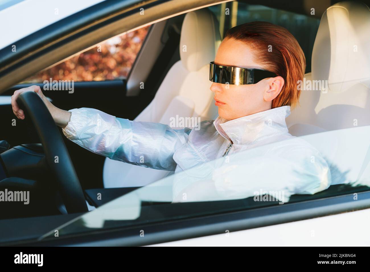 Moda cyberpunk ragazza guidare una supercar. Donna capelli rossi con occhiali futuristici siede accanto al volante dell'auto. Il concetto di tecnologia del futuro. Selectiv Foto Stock