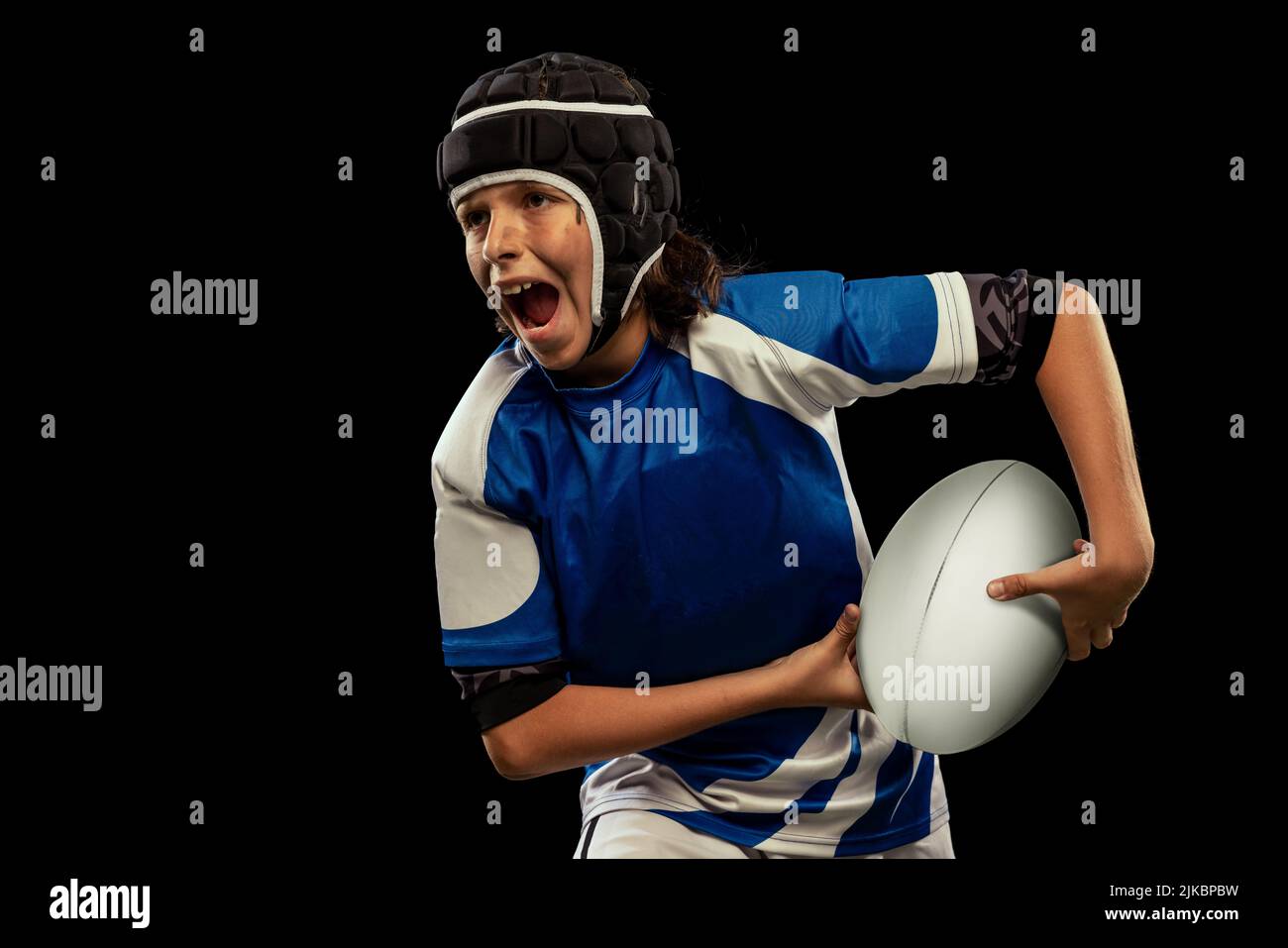Ritratto dinamico di ragazzo in età scolastica, giocatore di rugby maschile junior che pratica il calcio di rugby isolato su sfondo scuro con alluvione erba. Foto Stock
