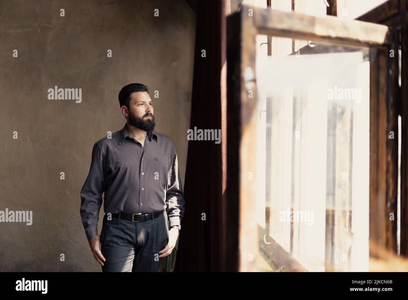 Un giovane uomo barbuto in un vestito guarda fuori la finestra Foto Stock