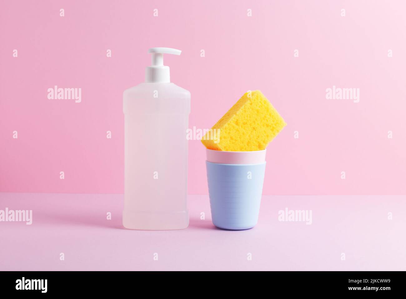 Vita morta creativa con stoviglie. Bottiglia bianca di liquido per lavastoviglie e spugna gialla per la pulizia della cucina in una pila di tazze di plastica. Vista frontale. Foto Stock