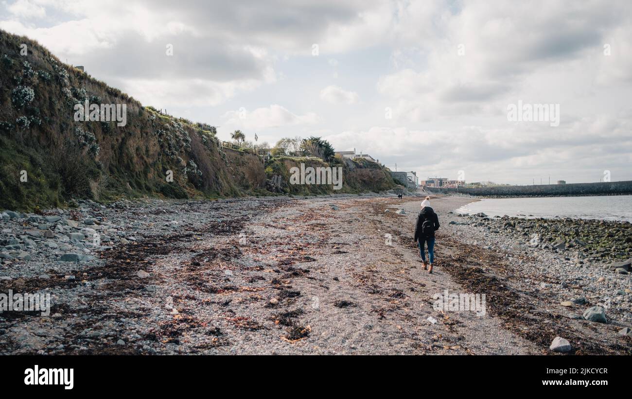Persona in giacca nera che cammina sulla spiaggia in Irlanda in una giornata fredda. La spiaggia è coperta di alghe. Foto Stock