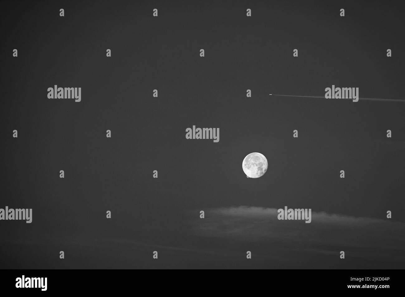 La luna piena rotonda nel cielo mattutino in bianco e nero, incorniciata in contrasto da un aereo con contrasti e una delicata nuvola morbida. Foto Stock