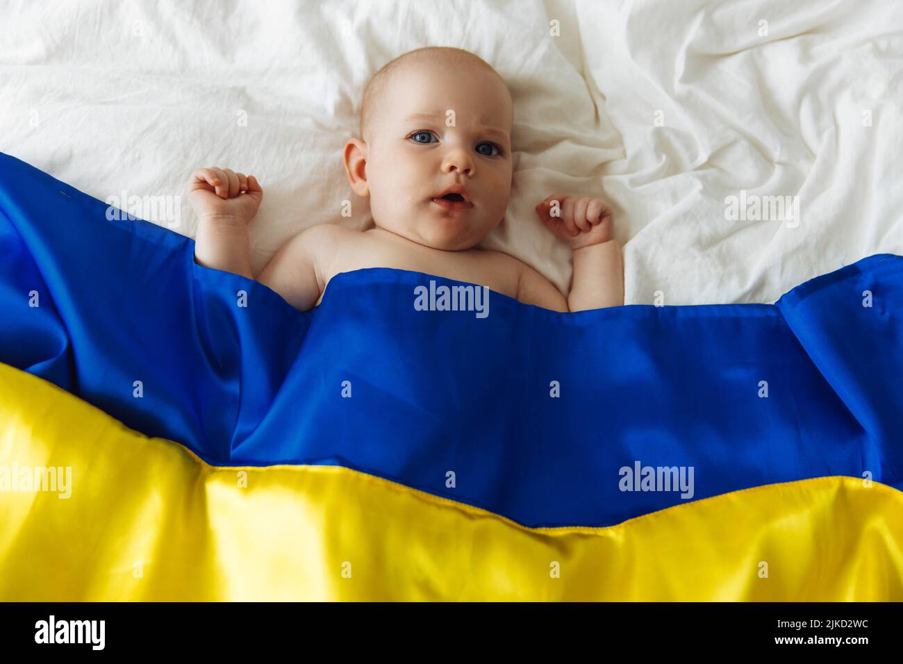 Ritratto di un bambino avvolto nella bandiera nazionale blu e gialla dell'Ucraina, sdraiato sul letto. Bambino con cuore bandiera Ucraina. Foto con posto per il testo. Foto Stock