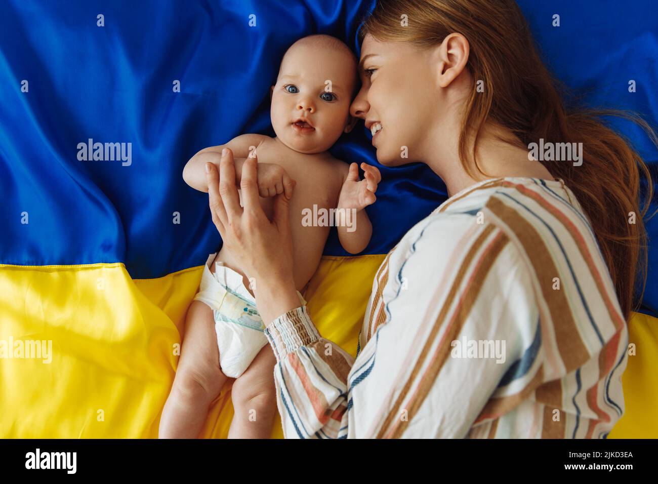 Amorevole madre premurosa con il suo neonato si trova sullo sfondo della bandiera Ucraina blu e gialla. La mamma bacia e abbraccia il bambino proteggendolo Foto Stock