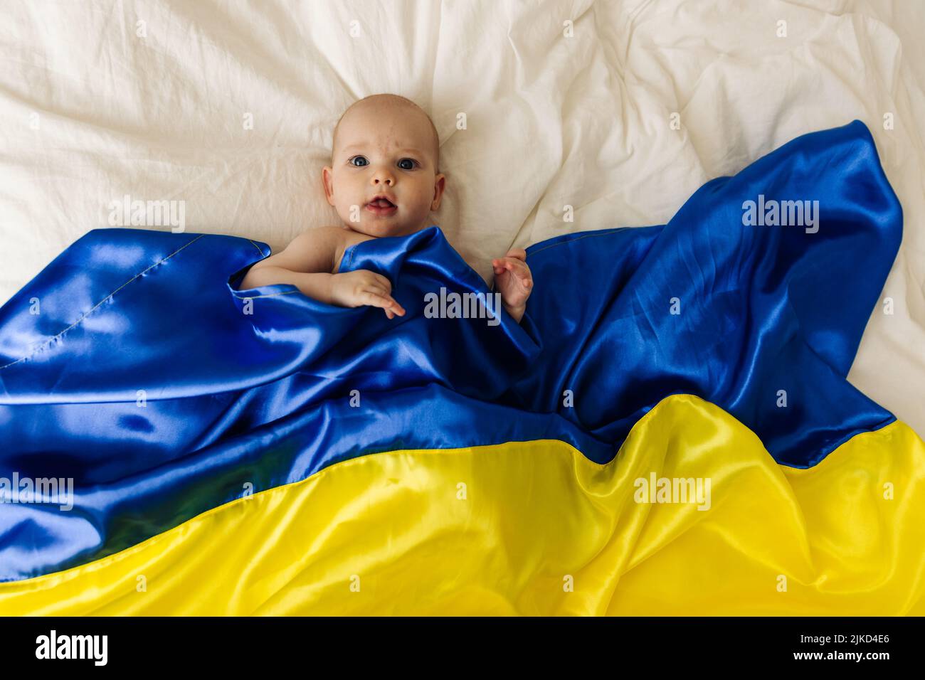 Ritratto di un bambino avvolto nella bandiera nazionale blu e gialla dell'Ucraina, sdraiato sul letto. Bambino con cuore bandiera Ucraina. Foto con posto per il testo. Foto Stock