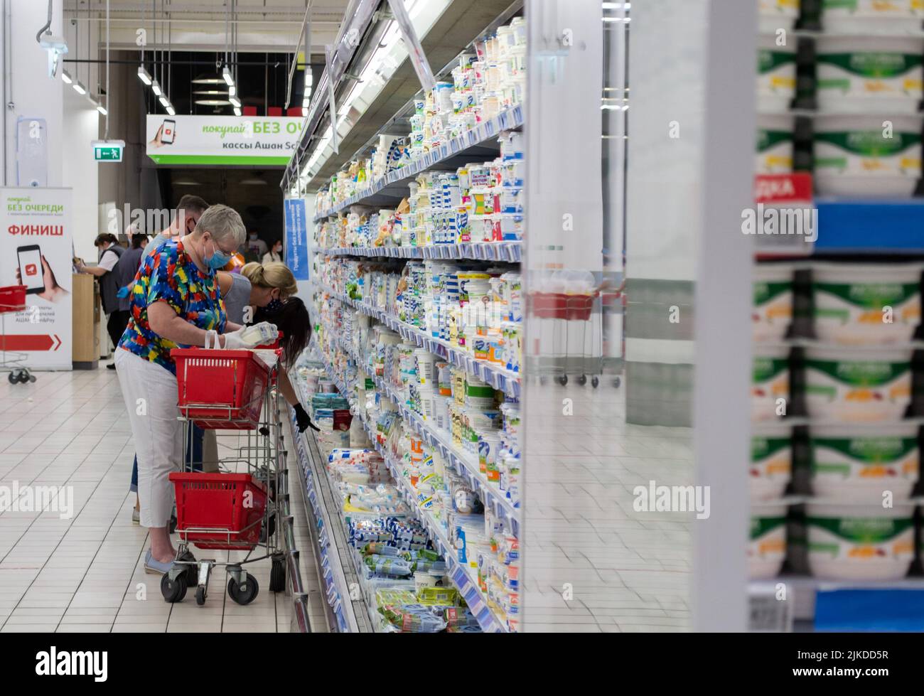 Mosca, Russia, Agosto 2020: Donna anziana in una maschera protettiva e guanti con cestini per lo shopping guarda il prodotto nel reparto caseario. Su un blu Foto Stock