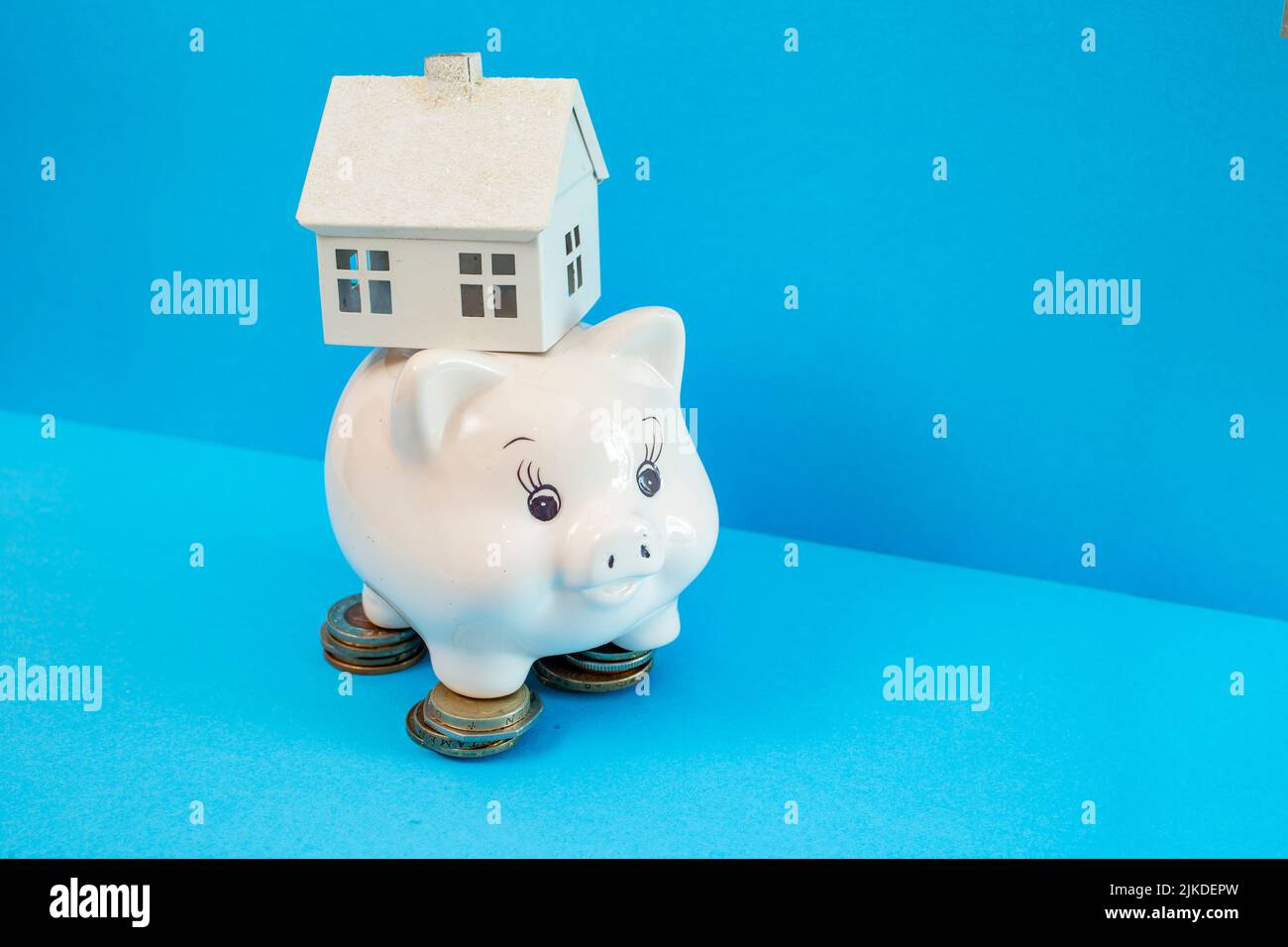 Porcellana bianca piggybank, in piedi su pile di monete con una casa modello sul retro, in primo piano isolato su sfondo blu Foto Stock